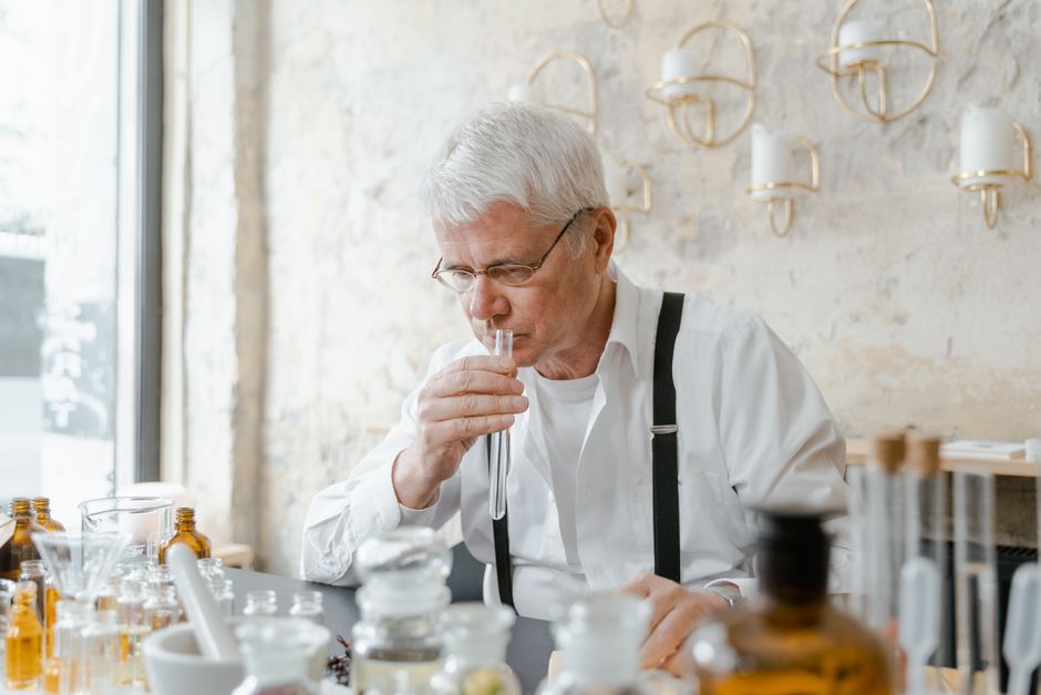 Senior man carefully smelling a test tube amidst laboratory glassware indicating a chemical experiment.